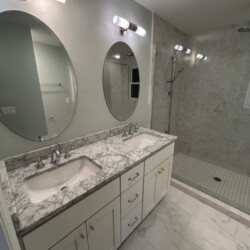 Modern bathroom with double sinks, marble countertop, oval mirrors, and a walk-in shower with glass door.