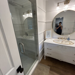 Bathroom with glass shower, round mirror above a white vanity, wood floor, and person taking a photo in the mirror.