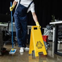 Person mopping office floor near a “Caution: Wet Floor” sign, with cleaning equipment and desks visible.
