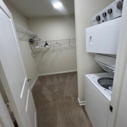 Walk-in closet with carpet, empty wire shelves, several hangers, and a stacked washer and dryer unit on the right.