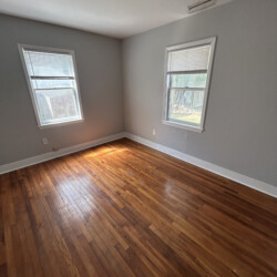 Empty room with two windows, light gray walls, wooden floor, and ceiling light fixture. Sunlight streams through one window.