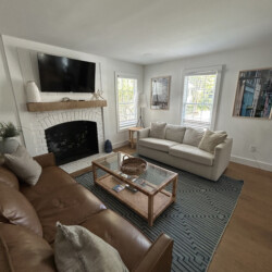 Living room with brown leather sofa, white loveseat, glass coffee table, fireplace, TV, and large windows.