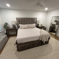 Bedroom with wicker bed frame, neutral bedding, two lamps on side tables, and a large mirror against the wall.