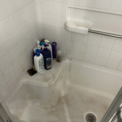 Shower with white tile walls, several bottles on corner ledge, and visible soap scum and grime on the floor.