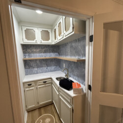 Small kitchenette with white cabinets, wood shelves, a sink, and a box on the counter; palm tree logo on the floor.