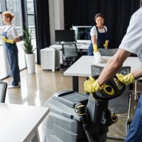 Three workers in uniforms clean an office; one uses a floor machine while others wipe surfaces and windows.