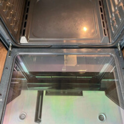 Open oven with clean interior and glass door, reflecting part of the kitchen and a person holding a phone.