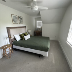 Minimalist bedroom with a green bedspread, two pillows, desk under window, ceiling fan, and neutral decor.