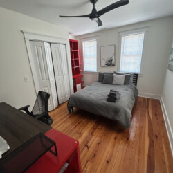 Bedroom with a bed, stacked towels, desk, chair, ceiling fan, and red shelves on a wooden floor.