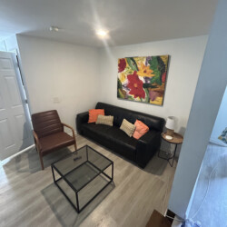 Small living room with a black sofa, glass coffee table, armchair, colorful painting, and wood flooring.