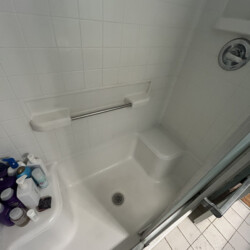 White tiled shower with a built-in seat, grab bar, assorted bottles, and a drain in the center of the floor.