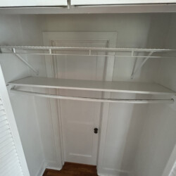 Empty white closet with wire shelf, hanging rod, and a door at the back, featuring wood floor and white walls.