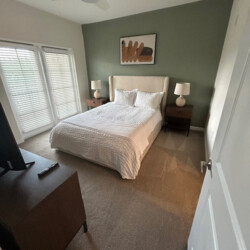 Bedroom with a neatly made bed, two nightstands, lamps, a dresser, TV, and a large window with blinds.